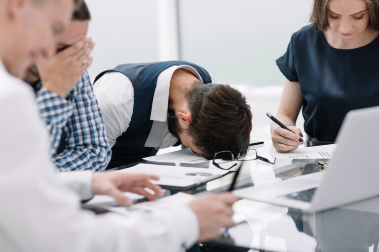 Professional employee experiencing meeting fatigue, laying his head on the table while another man covers his face with his hand and others discuss paperwork.