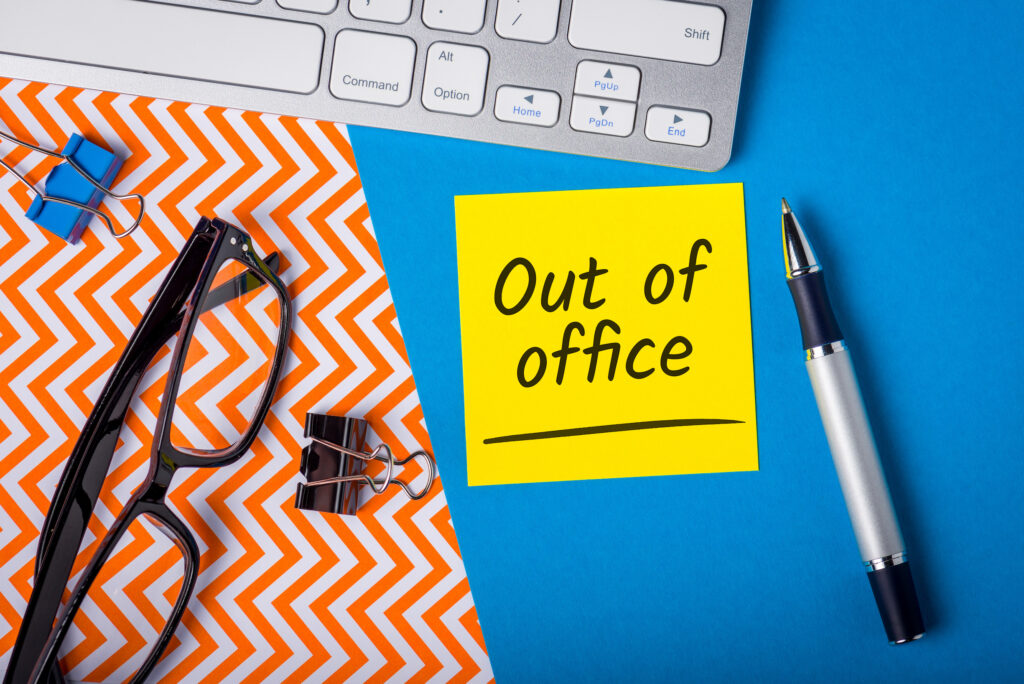 Sticky note with "out of office" written on it, laptop in background.